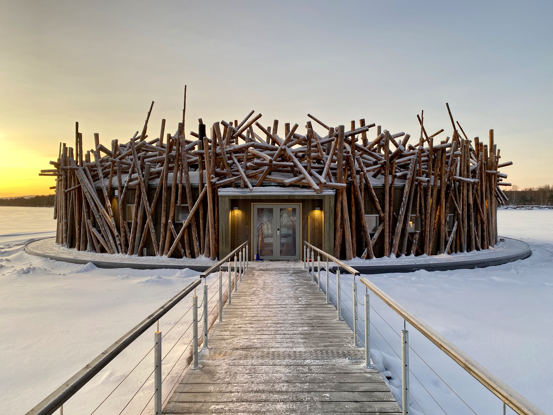 Circular timber lodge on a frozen lake at sunrise, connected by a wooden walkway